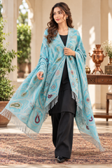 A woman models a light blue shawl with intricate multicolor paisley and floral patterns and white fringe, worn over a black outfit in an elegant outdoor courtyard setting.