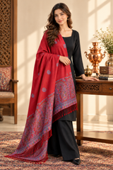 A woman models a red shawl with intricate blue and gold patterns and fringe, draped over a black outfit in a warm, traditional room setting.