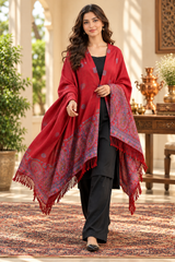A woman models a vibrant red shawl with intricate blue and gold patterns and fringed edges, worn over a black outfit in an elegant indoor setting with classical columns and warm lighting.