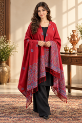 A woman models a rich red shawl with intricate blue and gold floral patterns and fringed edges, worn over a black outfit in an elegant indoor setting.