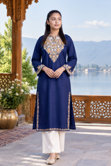 A woman stands outdoors on a stone terrace wearing a navy blue traditional kurta with ornate gold and silver embroidery around the neckline, cuffs, and hem, paired with white wide-leg pants. The background features a wooden pavilion, a large bronze pot, and a scenic lake and mountain landscape.