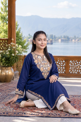 A woman sits cross-legged on a patterned rug on a wooden balcony, wearing a navy blue kurta with intricate gold embroidery around the neckline and hem, paired with white pants. The background features a scenic view of a lake and mountains.
