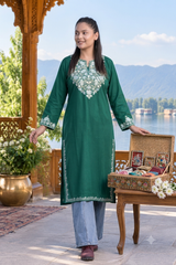 A woman wearing a green long kurta with white floral embroidery stands on an outdoor porch beside a wooden table holding a decorative box. The background shows a scenic lake and mountains.