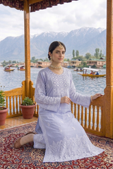 A woman wearing a light lavender embroidered traditional outfit kneels on a patterned rug on a wooden balcony, with a scenic lake and mountains in the background.