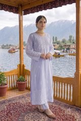 A woman stands on a wooden balcony wearing a light lavender embroidered kurta and matching trousers. The background features a scenic lake with boats and mountains under a cloudy sky.