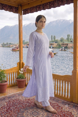A woman models a light lavender purple embroidered long kurta with matching trousers on a wooden boat porch with a scenic mountain and lake background.
