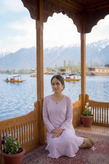 A woman in a light lavender traditional outfit sits on a wooden balcony overlooking a lake with mountains in the background.