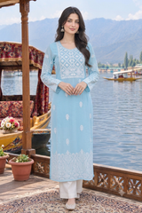 A woman stands on a wooden dock by a lake wearing a light blue embroidered kurta with white patterns and white pants. Boats and mountains are visible in the background.