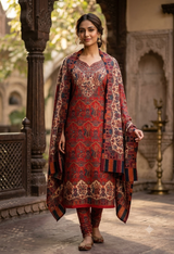 A woman modeling a traditional Indian salwar kameez suit in a courtyard setting. The outfit features a red kurta with intricate gold and blue patterns, matching red trousers, and a coordinating red dupatta with blue and gold borders.