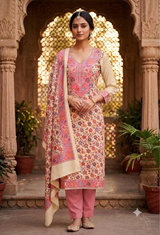 A woman stands in an ornate stone courtyard wearing a traditional Indian salwar kameez suit. The outfit features a pink and beige floral printed kurta with a beige inner collar, pink trousers, and a matching pink dupatta with a beige border.