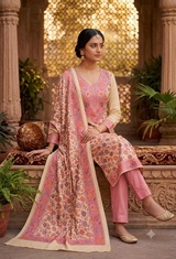 A woman models a traditional Indian salwar kameez suit set in a beautifully carved courtyard. The set features a light beige kameez jacket and trousers with pink floral patterns, paired with solid pink pants and a matching pink dupatta with a beige border.