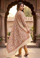 A woman wearing a traditional Indian salwar kameez outfit with a beige floral print kurta and dupatta, paired with mustard yellow pants, standing in front of an ornate stone archway.