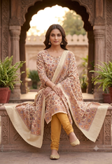 A woman models a traditional Indian salwar kameez suit set consisting of a beige and brown floral print kurta, matching dupatta, and mustard yellow trousers, seated on a stone bench in front of an ornate archway.