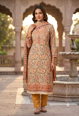 A woman stands in a courtyard with arches and a fountain, wearing a traditional printed salwar kameez outfit. The outfit has a warm beige base with intricate multicolor floral embroidery and a matching dupatta, paired with mustard yellow trousers.