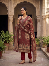 A woman stands in a courtyard wearing a traditional Indian outfit consisting of a maroon kurta with gold and beige patterns, matching maroon pants, and a patterned dupatta draped over her shoulder.