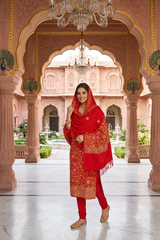 A woman modeling a traditional red Indian salwar kameez suit with intricate gold embroidery and a matching red dupatta, standing in a grand pink stone courtyard with arches and chandeliers.