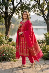 A woman stands outdoors by a lake wearing a bright pink traditional outfit with ornate gold embroidery and a matching embroidered dupatta.