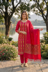 A woman stands outdoors by a lake wearing a bright red traditional outfit with gold embroidery, matching pants, and a large red embroidered dupatta draped over one arm. Trees, flowers, and a stone path are visible in the background.