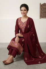 A woman seated indoors wearing a deep maroon traditional outfit with pink floral embroidery and a matching draped stole against a plain light wall.