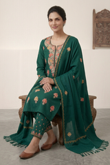A woman seated on a wooden chair wearing a dark green traditional outfit with floral embroidery and a matching draped scarf, photographed indoors against a light neutral wall.
