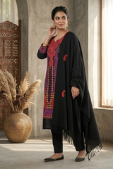 A woman models a black kurta set with a red and purple embroidered tunic and a matching black fringed shawl, standing in a rustic room with a large clay pot of dried grass.