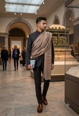 A man in a black suit and brown dress shoes walks through a museum gallery, draped with a beige shawl over his shoulder.