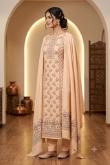 A woman models a traditional Indian salwar kameez suit in a warm beige/peach color with intricate dark brown embroidery. The set includes a long kurta, matching trousers, and a draped dupatta, shown in a beautifully lit interior with arches and wooden floors.