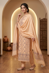 A woman models a traditional Indian salwar kameez suit in a warm beige and peach color palette, featuring intricate embroidery on the kurta and a matching dupatta, set against an elegant arched hallway with warm lighting.
