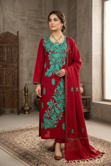 A woman models a traditional Indian salwar kameez suit in a rustic indoor setting. The outfit consists of a deep red kurta with vibrant teal green floral embroidery, matching red trousers, and a coordinating red dupatta.