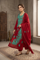 A woman models a traditional Indian salwar kameez suit in a rustic indoor setting. The outfit consists of a deep red kurta and trousers with vibrant green floral embroidery, paired with a matching red dupatta featuring green embroidered details.