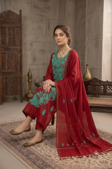 A woman seated indoors wearing a traditional red salwar kameez outfit with a teal embroidered kurta and a matching red dupatta draped over her lap, set against a rustic carved wooden furniture backdrop.
