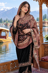 A woman models a traditional outfit featuring a black velvet kurta with gold embroidery and a large brown and gold patterned shawl, set against a scenic background of a lake, boats, and mountains.