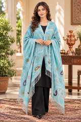 A woman stands in an elegant indoor courtyard setting wearing a light blue shawl with intricate multicolor paisley and floral patterns, draped over a black outfit. The shawl features white fringe along the edges.