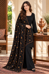 A woman models a black three-piece salwar kameez suit with a matching printed dupatta in a traditional indoor setting. The outfit features a black kurta, wide-leg trousers, and a black dupatta decorated with gold floral patterns.