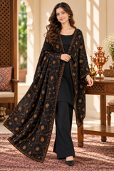 A woman models a black three-piece traditional outfit with a matching long open-front shawl draped over her shoulders, set in a warm, traditional interior with wooden furniture and a patterned rug.