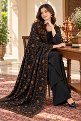 A woman models a black three-piece ethnic salwar suit set with a brown floral print dupatta, sitting on a wooden chair in an elegant indoor setting.