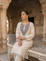 Full-length view of a model seated in an ornate marble corridor wearing a traditional off-white Aari embroidered phiran kurta.