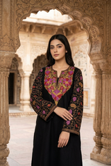 Pakistani model wearing a black Kashmiri phiran kurta with colorful machine Sozni embroidery on the neck and sleeves, seated in an ornate palace courtyard.