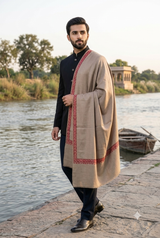 Handsome bearded model in a black sherwani wearing a natural beige Kashmiri shawl with a rust embroidered border by a scenic riverside.