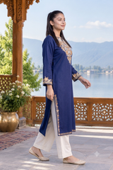 A woman modeling a navy blue kurta with gold embroidery and white pants, standing on a wooden balcony with a scenic lake and mountains in the background.