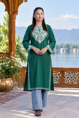 A woman stands on a balcony wearing a dark green kurta with white floral embroidery on the chest and cuffs, paired with light blue jeans. The background features a scenic lake and mountains under a clear blue sky.