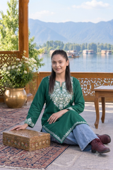A woman sits on a patterned rug in a wooden pavilion with a scenic lake and mountains in the background, wearing a green embroidered kurta with light blue jeans and brown shoes, next to a decorative wooden box.