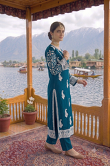 A woman models a teal blue and white embroidered traditional outfit with matching pants on a wooden balcony overlooking a scenic lake and mountains.