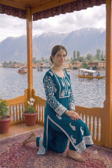 A woman kneels on a patterned rug on a wooden porch, wearing a teal blue and white embroidered traditional outfit. In the background, a scenic lake with boats and mountains is visible.
