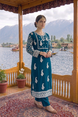 A woman models a teal blue and white embroidered traditional outfit on a wooden boat porch with a scenic mountain lake background.