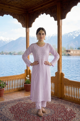A woman stands on a wooden balcony wearing a light pink embroidered kurta and matching pants, with a scenic lake and snow-capped mountains in the background.