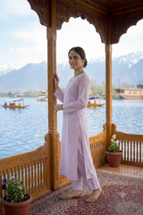 A woman wearing a light lavender embroidered kurta and matching trousers stands on a wooden boat platform with a scenic lake and mountains in the background.