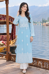 A woman models a light blue long kurta with white embroidery, paired with white pants, standing on a wooden dock with a scenic lake and mountains in the background.