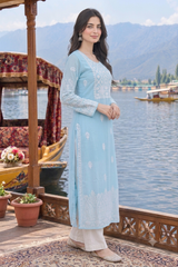 A woman stands on a wooden dock by a lake wearing a light blue embroidered kurta with white pants. Boats and mountains are visible in the background.