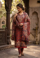 A woman wearing a traditional Indian salwar kameez suit in deep red and gold with intricate patterns, standing in an outdoor courtyard with stone pillars and arches.